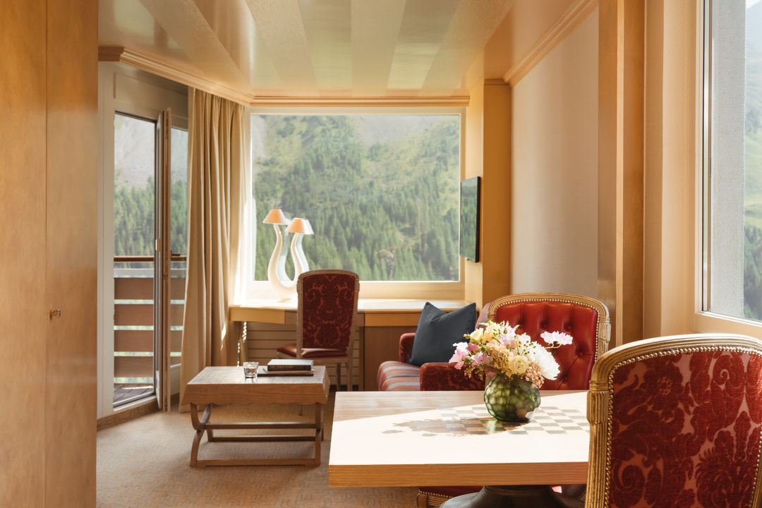 Heller Wohnbereich eines Hotels mit rot gepolsterten Sesseln, einem Holztisch mit Blumenarrangement, Schreibtisch und grossen Fenstern mit Panoramablick auf die Alpenlandschaft.