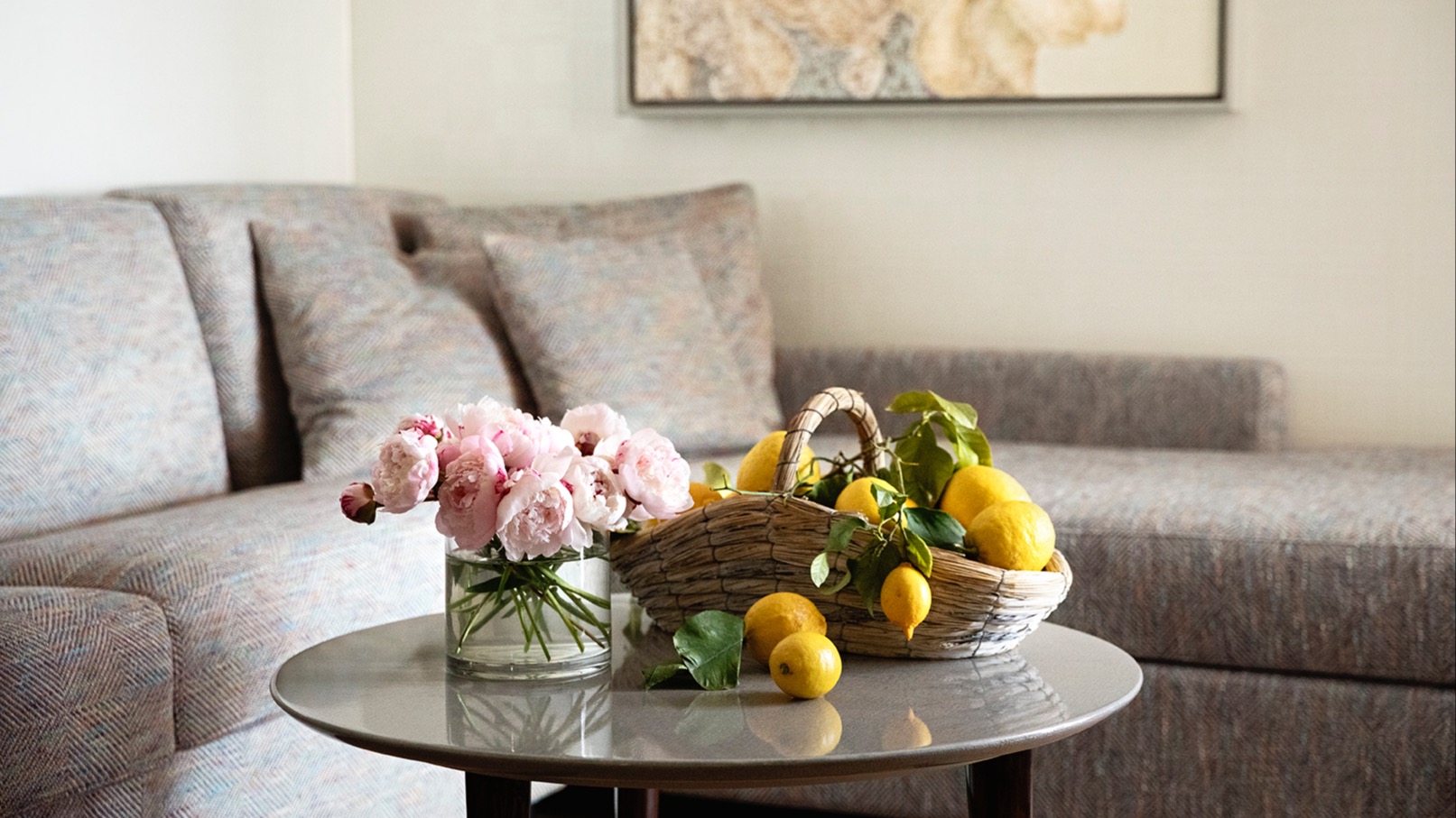 Nahaufnahme eines stilvollen Couchtischs mit frischem Zitronenkorb und rosa Pfingstrosen vor einem strukturierten Sofa und floraler Wandkunst.