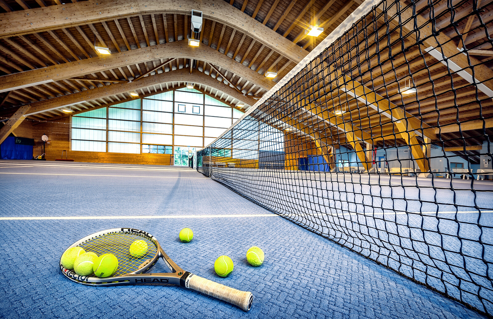 Indoor tennis court with wooden arched ceiling, net across center, tennis racket and yellow balls on blue court surface.