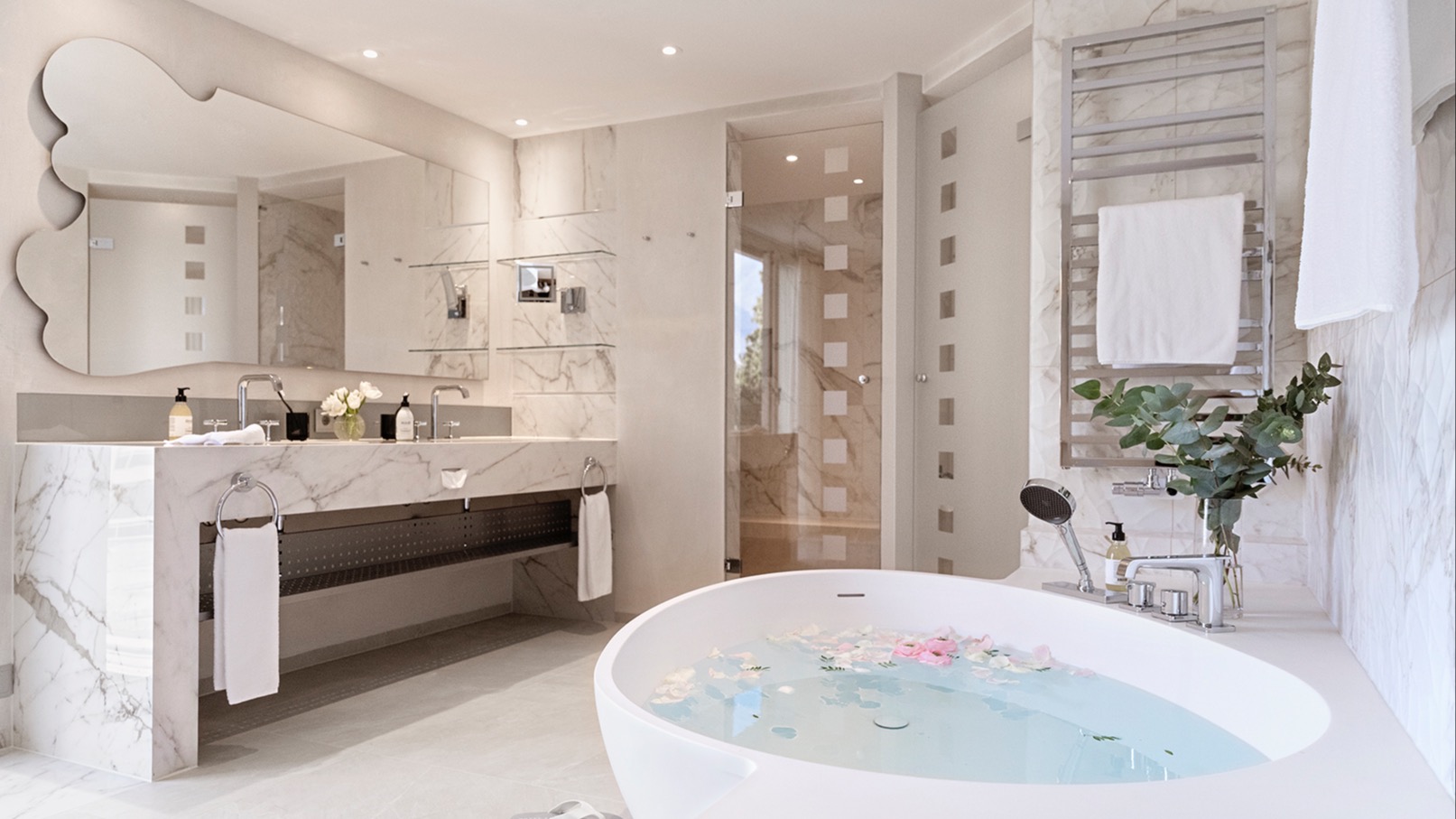 Modern designer bathroom with freestanding bathtub filled with petals, marble double sink vanity, mirrored wall design, and stylish fixtures.