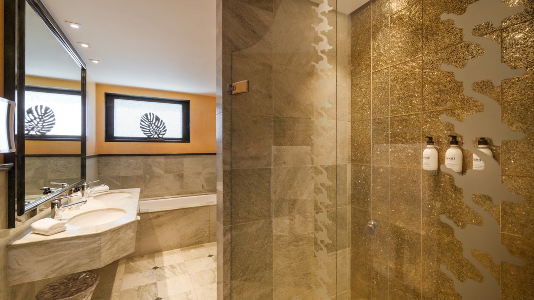 Luxurious bathroom featuring gold-tiled mosaic shower, natural stone flooring, marble double vanity, mirror, and decorative windows with leaf patterns.