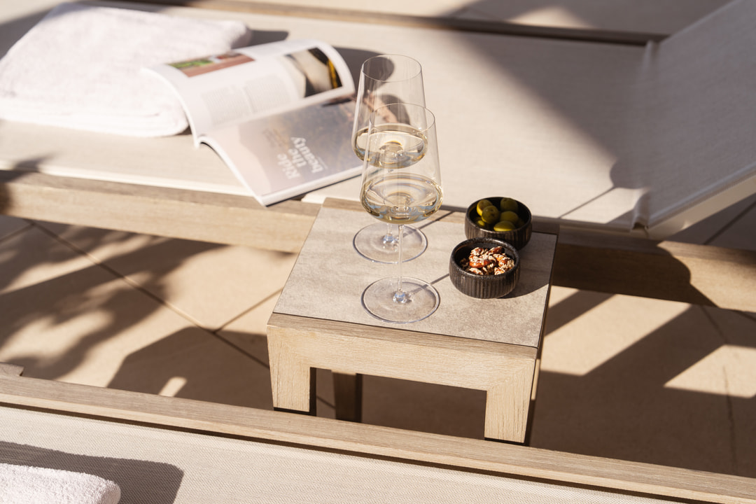 Light wooden side table with two wine glasses, small bowls of snacks, and an open magazine in bright sunlight.