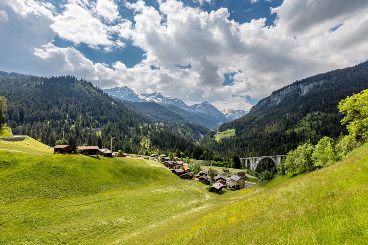 Schweizer Alpendorf mit grünen Wiesen, Holzchalets, Steinbogenbrücke, bewaldeten Bergen und schneebedeckten Gipfeln.