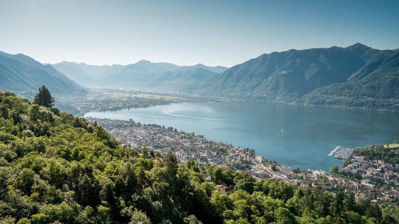 Panoramic view over Locarno and Lake Maggiore with the town, marina, and forested mountains in the background.