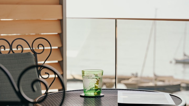 Detailaufnahme eines Balkons mit Metalltisch, grünem Trinkglas, Tablet und Blick auf den Hafen mit Segelbooten.