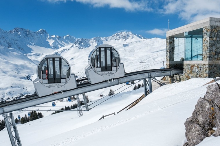 Moderne Seilbahnstation mit runden Gondeln vor schneebedeckten Alpengipfeln und blauem Himmel
