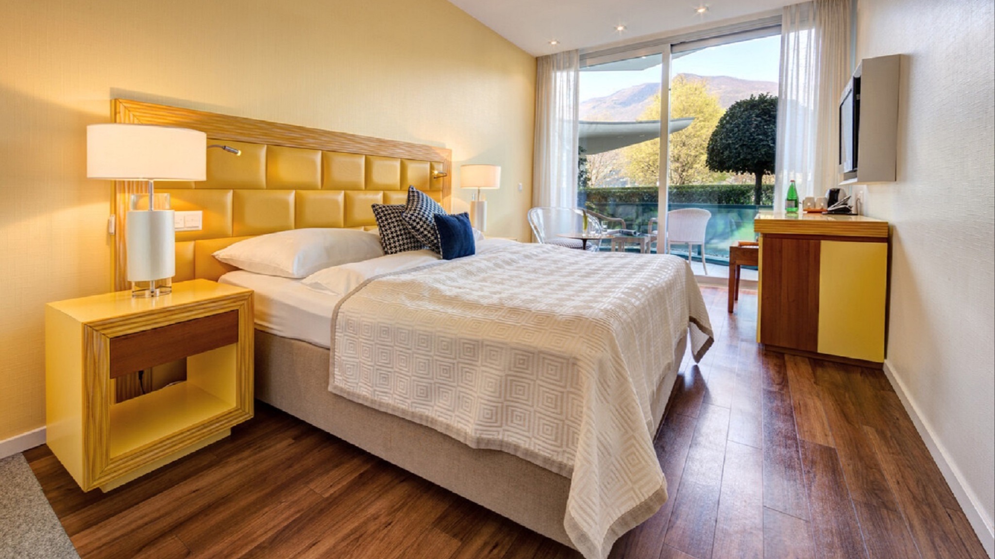 Modern hotel room with yellow wall, large double bed, textured bedspread, panoramic windows, access to terrace with garden view, and designer chairs.