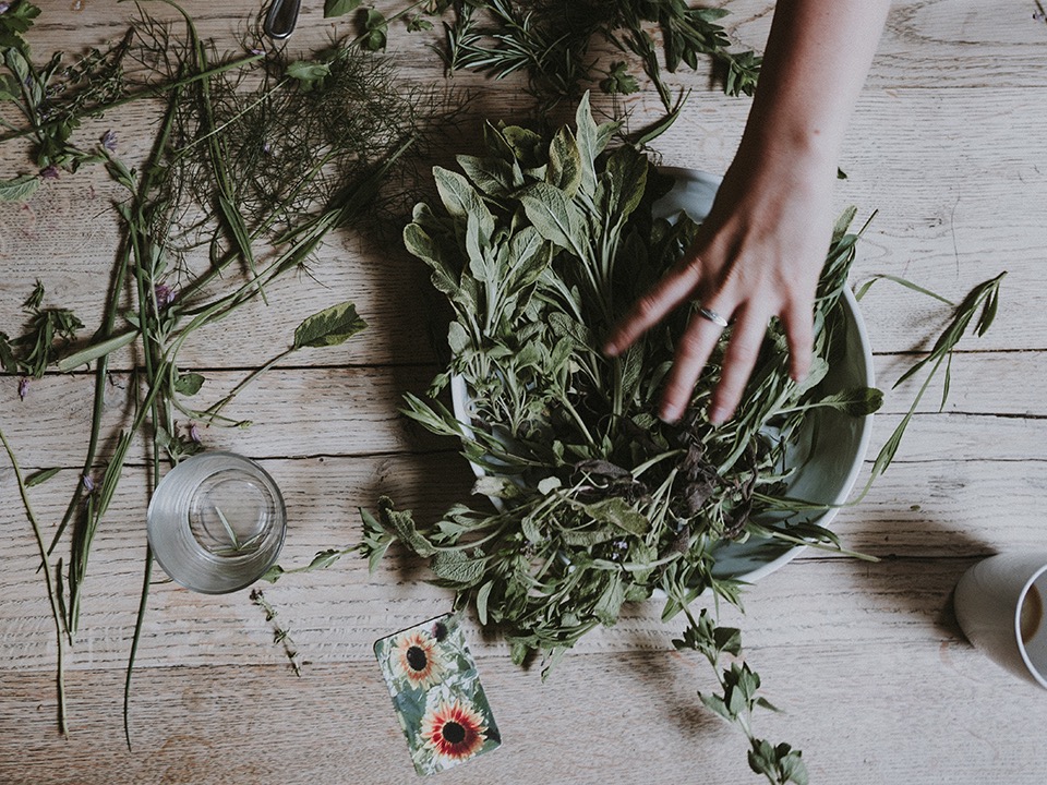Hand arranging fresh herbs and greenery in a bowl on a rustic wooden table with scattered stems and flowers nearby.
