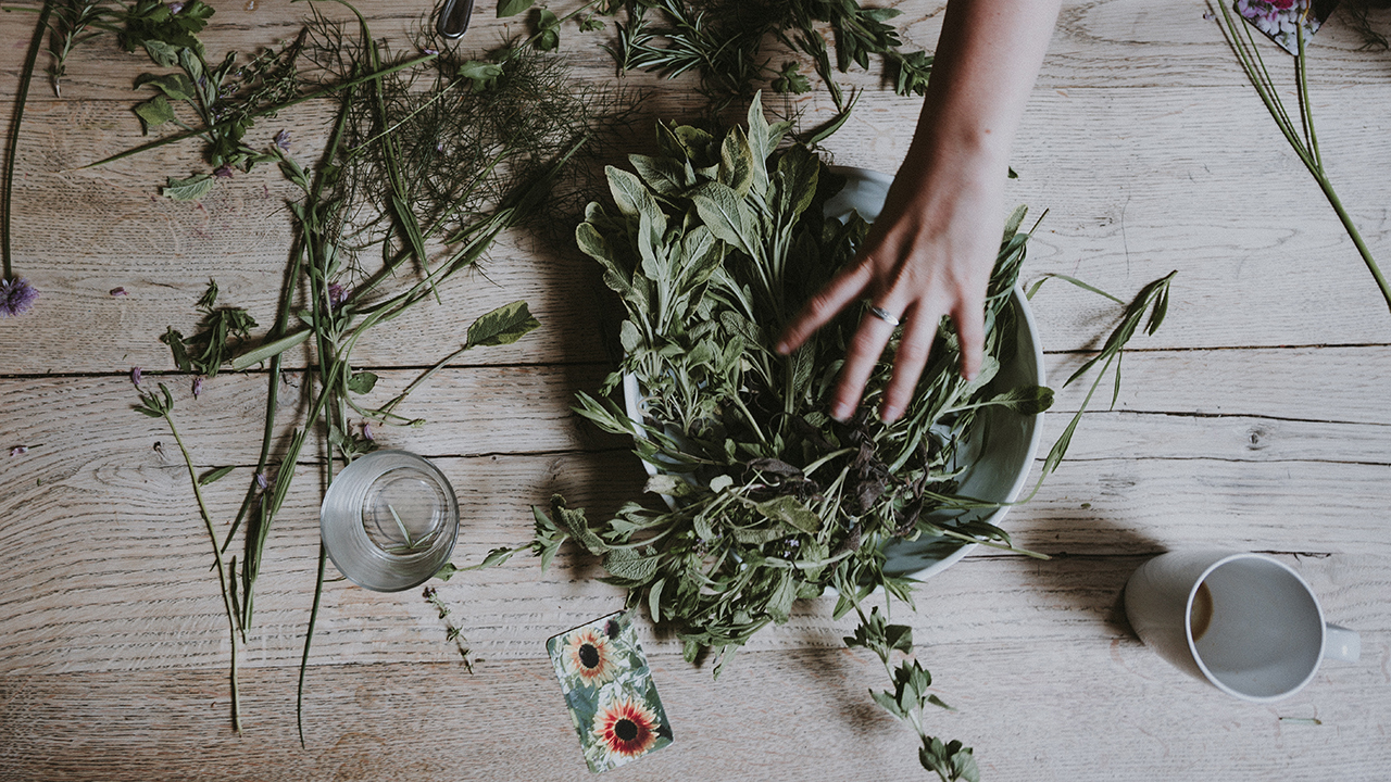 Hand arranging fresh herbs and greenery in a bowl on a rustic wooden table with scattered stems and flowers nearby.