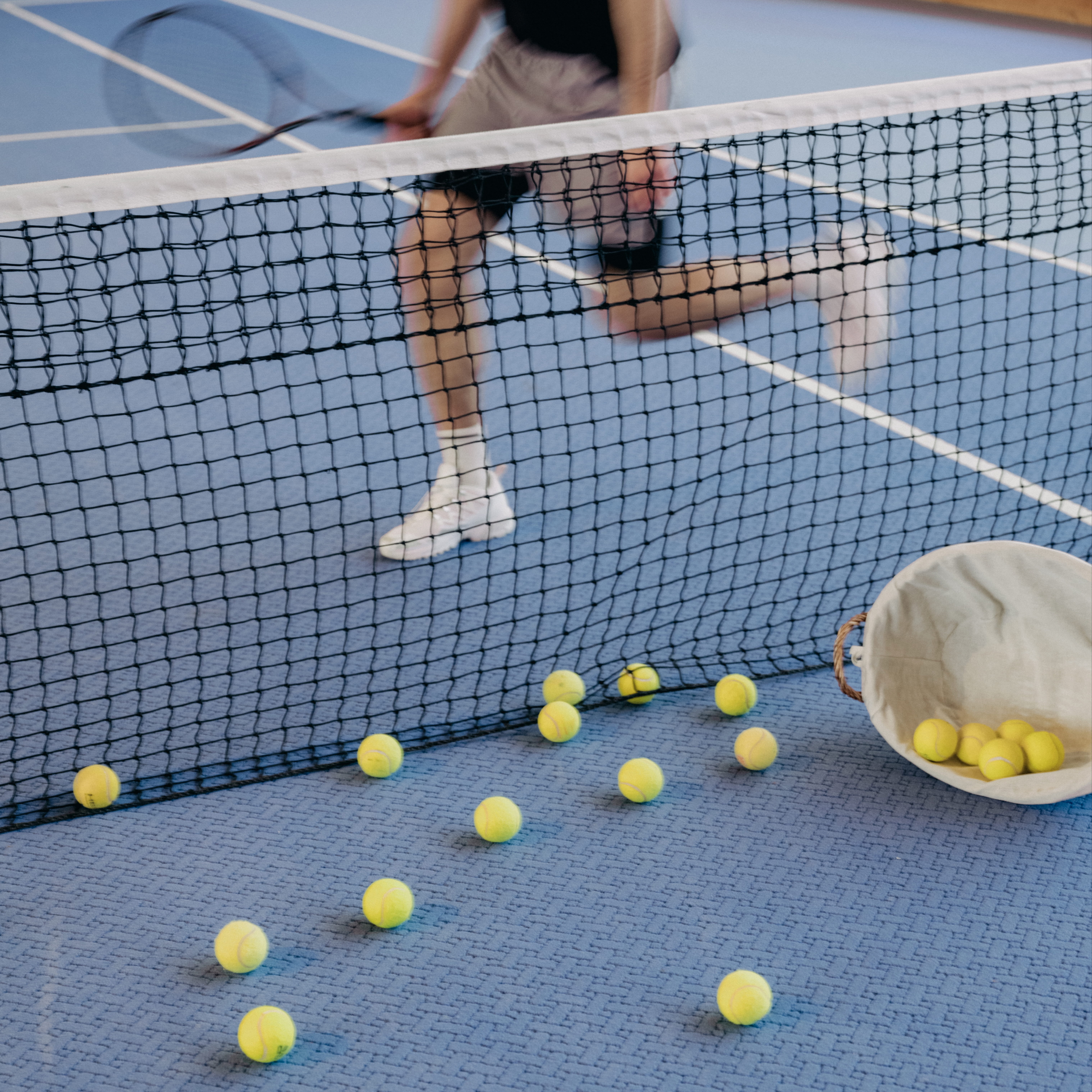 Tennis player holding racket behind net on blue court with scattered yellow tennis balls and basket.