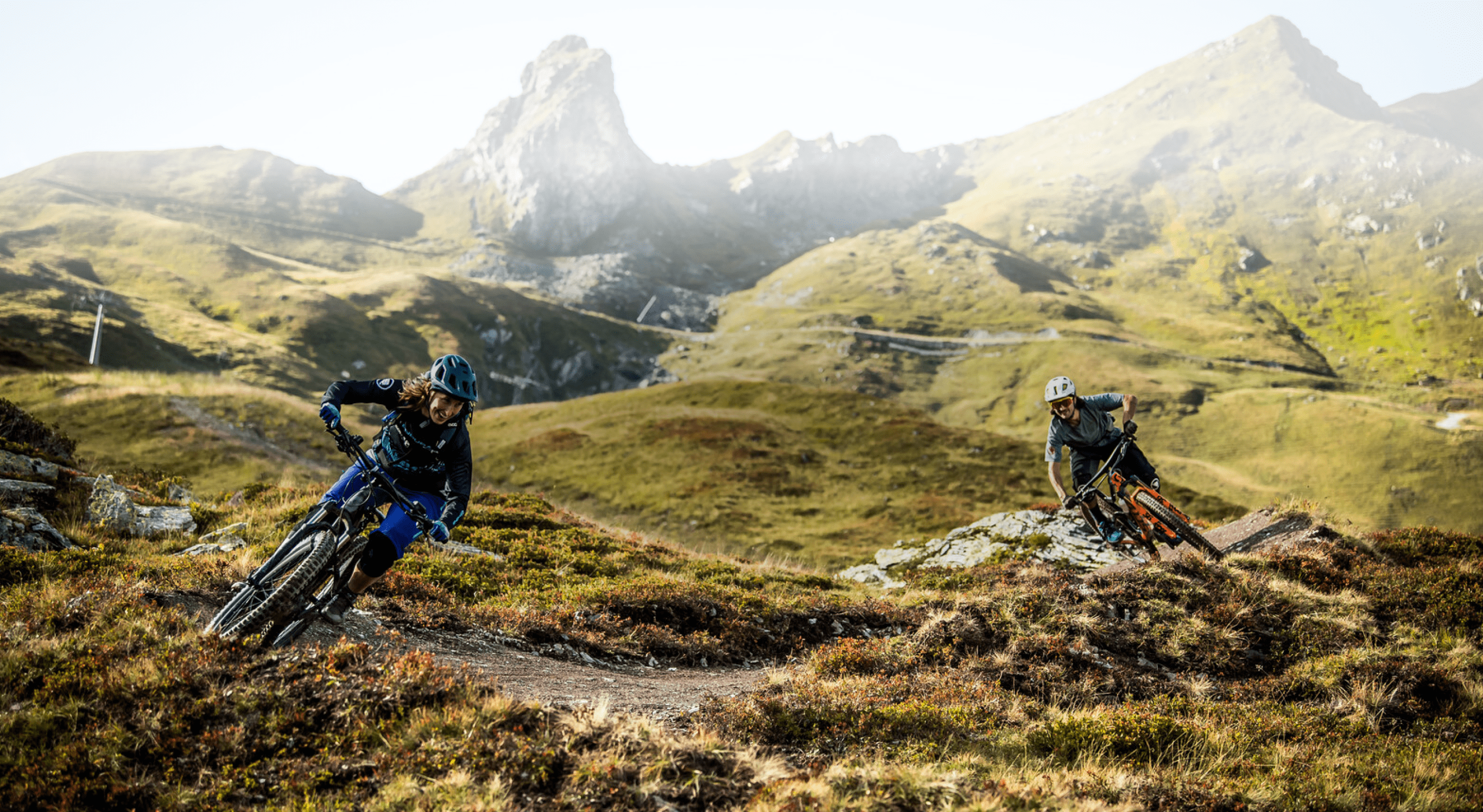 Zwei Mountainbiker fahren auf einem alpinen Trail mit Bergpanorama im Hintergrund.