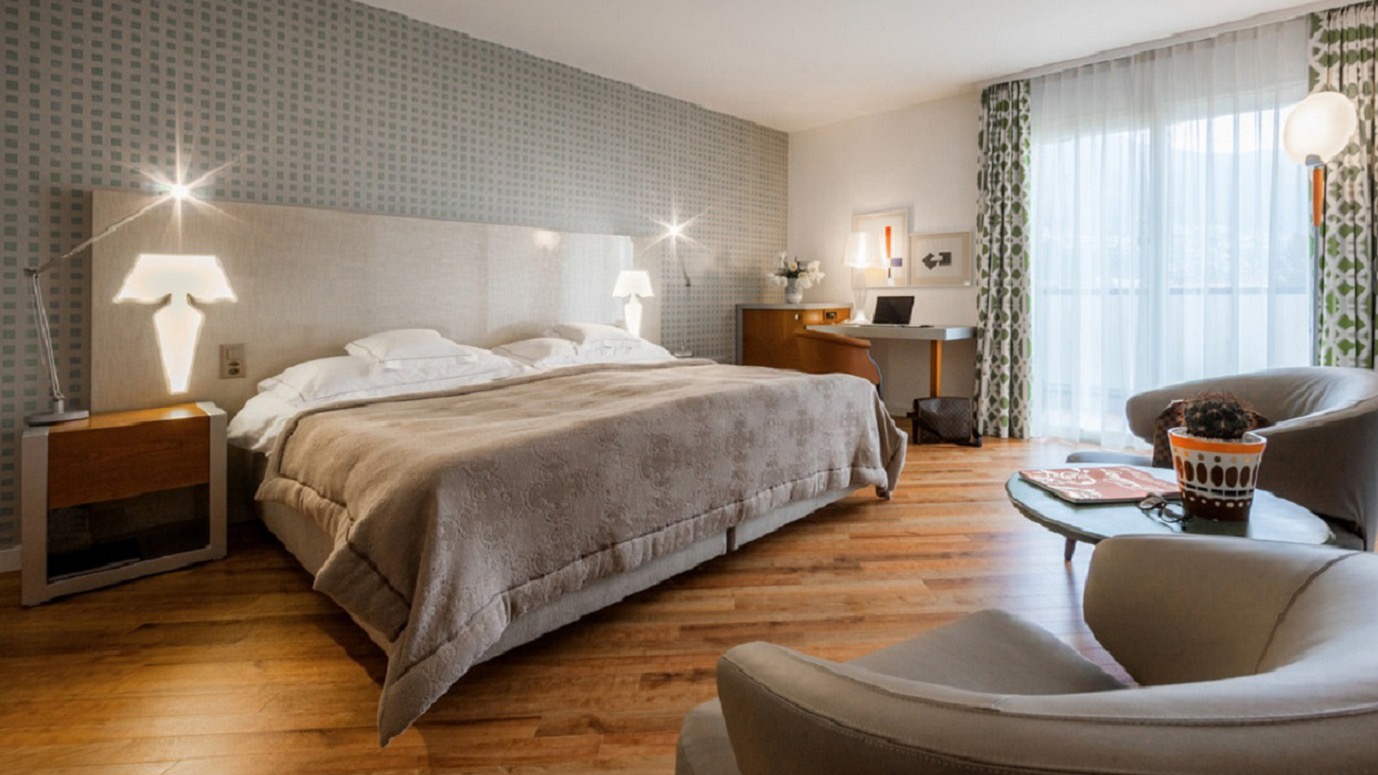 Modernes Hotelzimmer mit großem Doppelbett, gemütlicher Sitzecke, Schreibtisch, Holzboden, grafischer Akzentwand und Balkon mit Tageslicht.