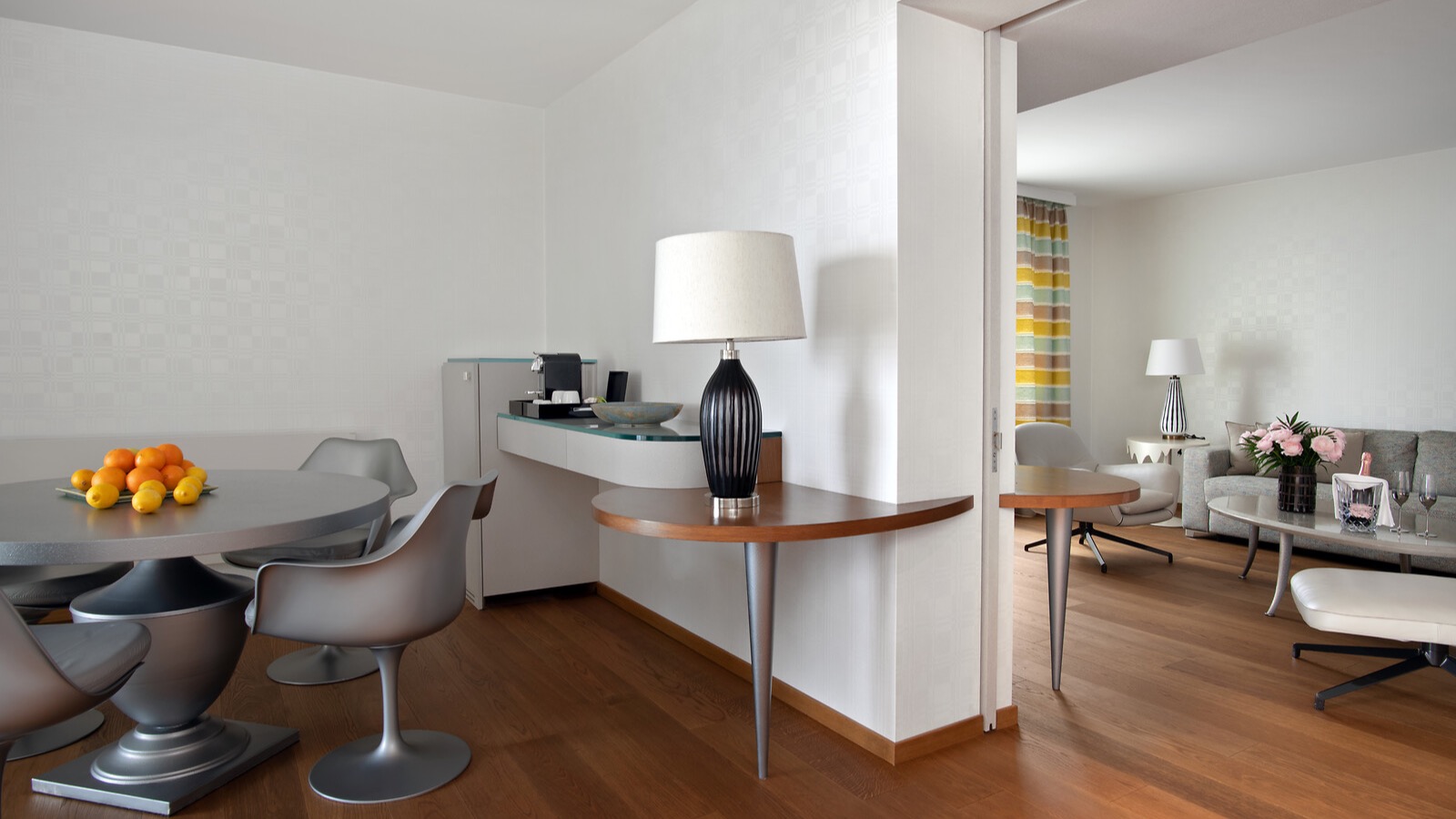 Contemporary dining area with round table, designer chairs, fruit bowl centerpiece, and adjacent living room with hardwood flooring and minimalist decor.