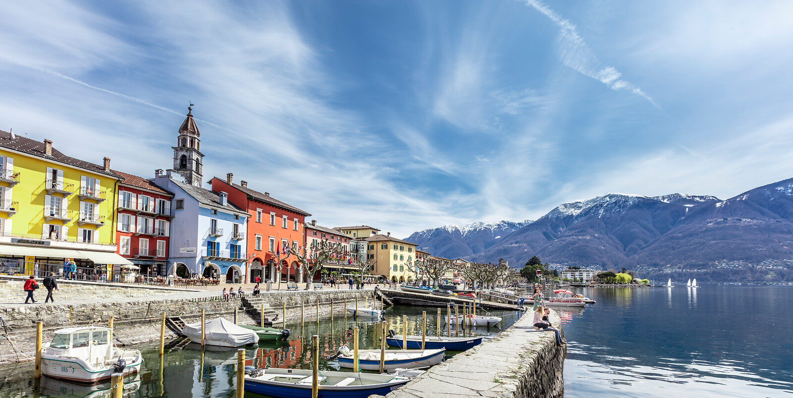 Bunte Häuser am Seeufer mit Booten, Kirchturm und schneebedeckten Bergen im Hintergrund unter blauem Himmel.