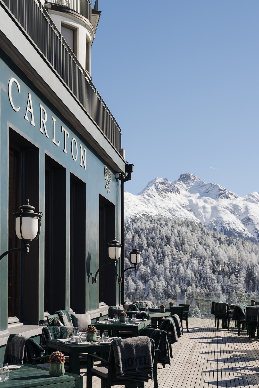 Carlton Hotel terrace with outdoor seating and snow-covered mountain peaks in the background under a clear blue sky.