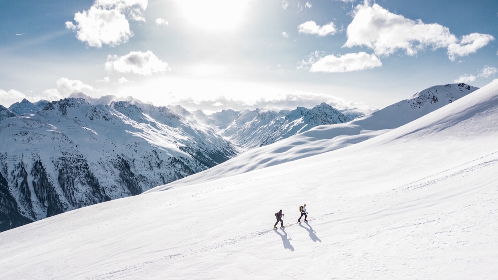 Two skiers ascending a snowy mountain slope with snow-covered peaks and blue sky with white clouds in the background.