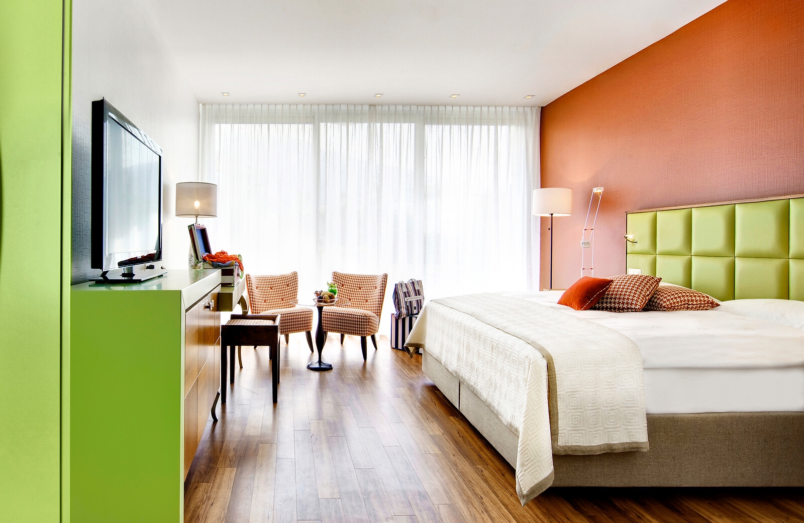 Farbintensives Hotelzimmer mit grünem Schrank, orangefarbener Wand, gelb-grünem Kopfteil, Parkettboden und Panoramafenster mit Sitzbereich.