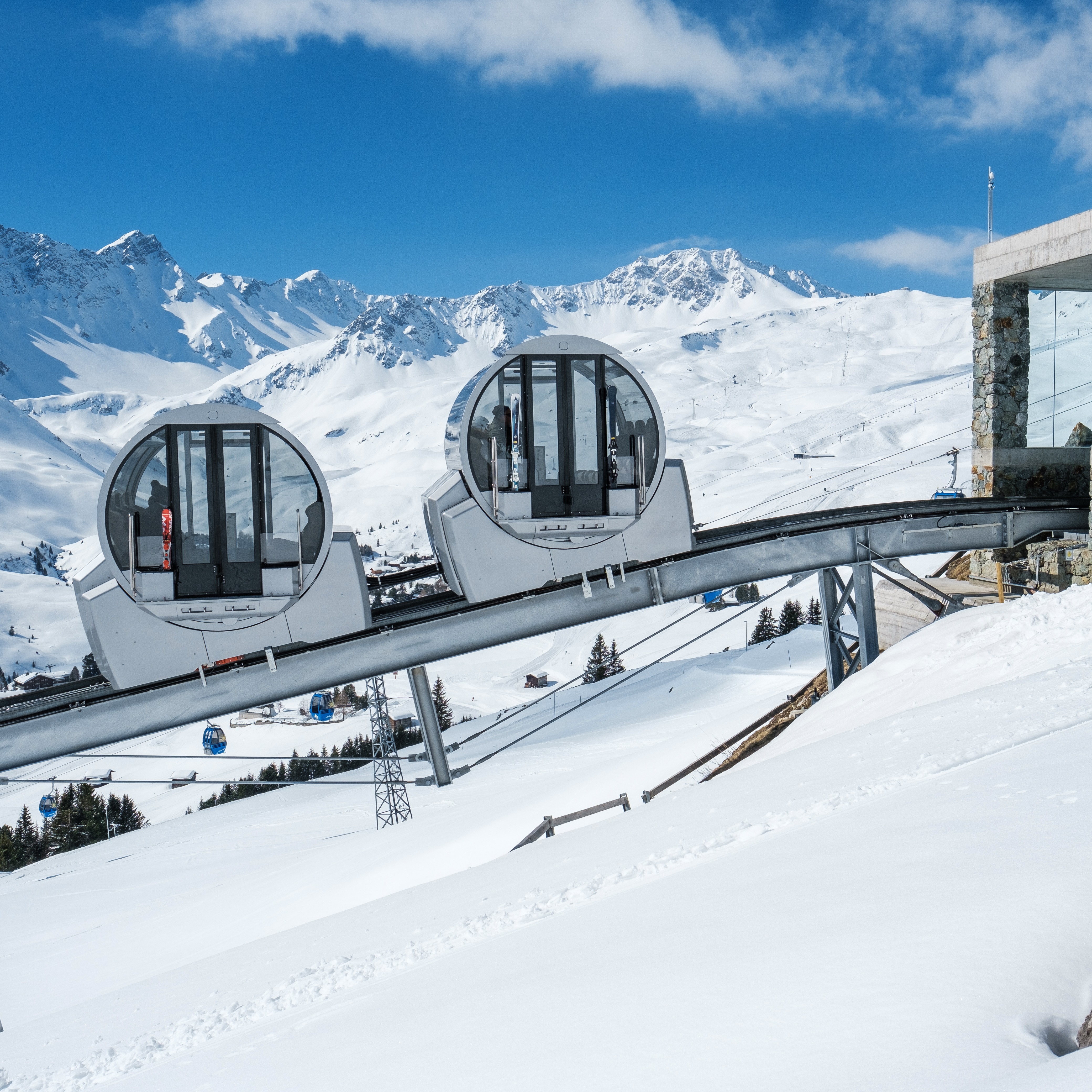 Modern circular ski gondolas with glass windows on cables ascending snowy mountain slopes under blue sky with peaks behind.