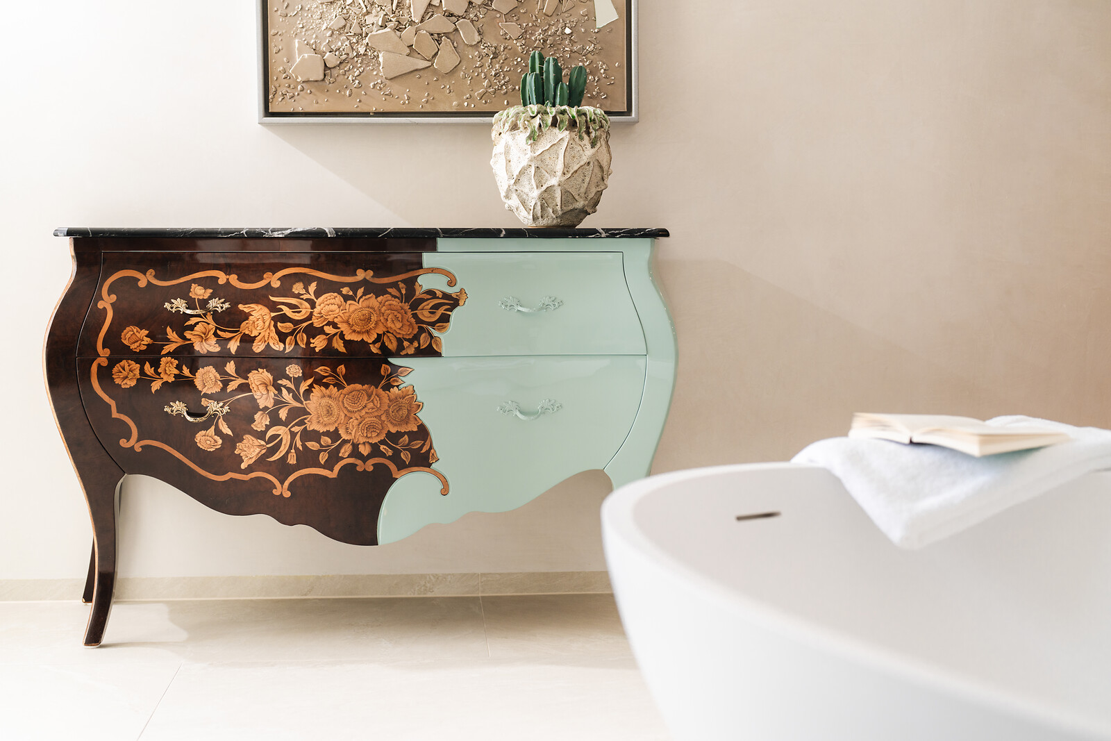 Close-up of an artistic designer dresser with floral inlay, half-painted in mint green, beside a freestanding bathtub with towel and open book.