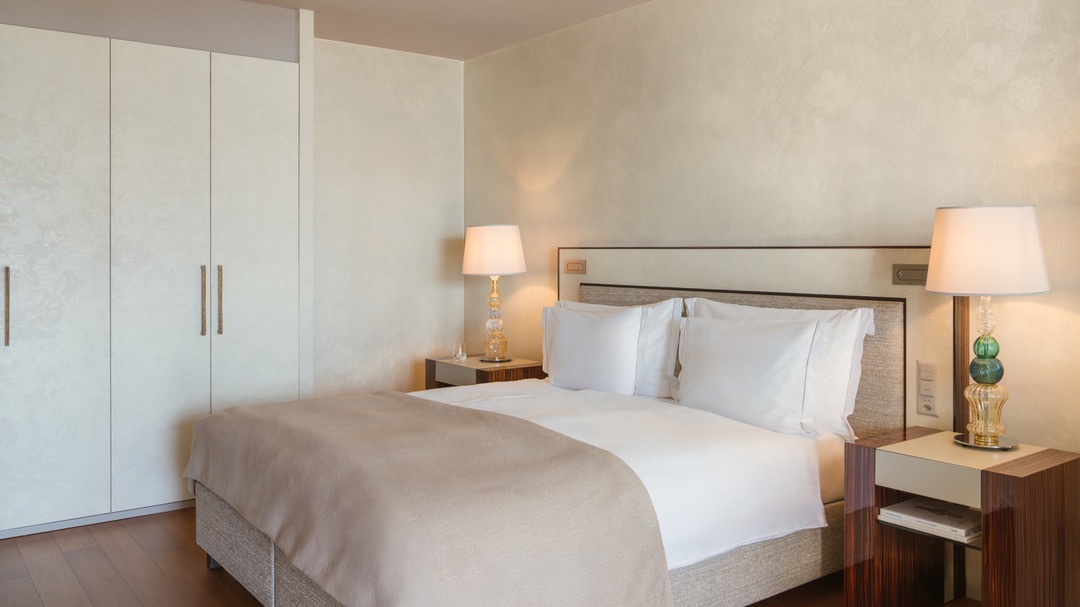 Modern bedroom with beige double bed, white pillows, two bedside tables with decorative lamps, built-in wardrobe, and warm wooden flooring.