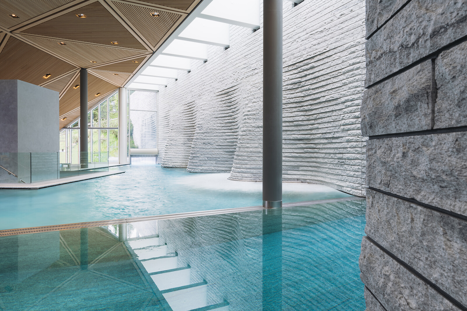 Moderner Innenpool mit türkisfarbenem Wasser, geschwungenen Steinwänden und geometrischer Holzdecke.