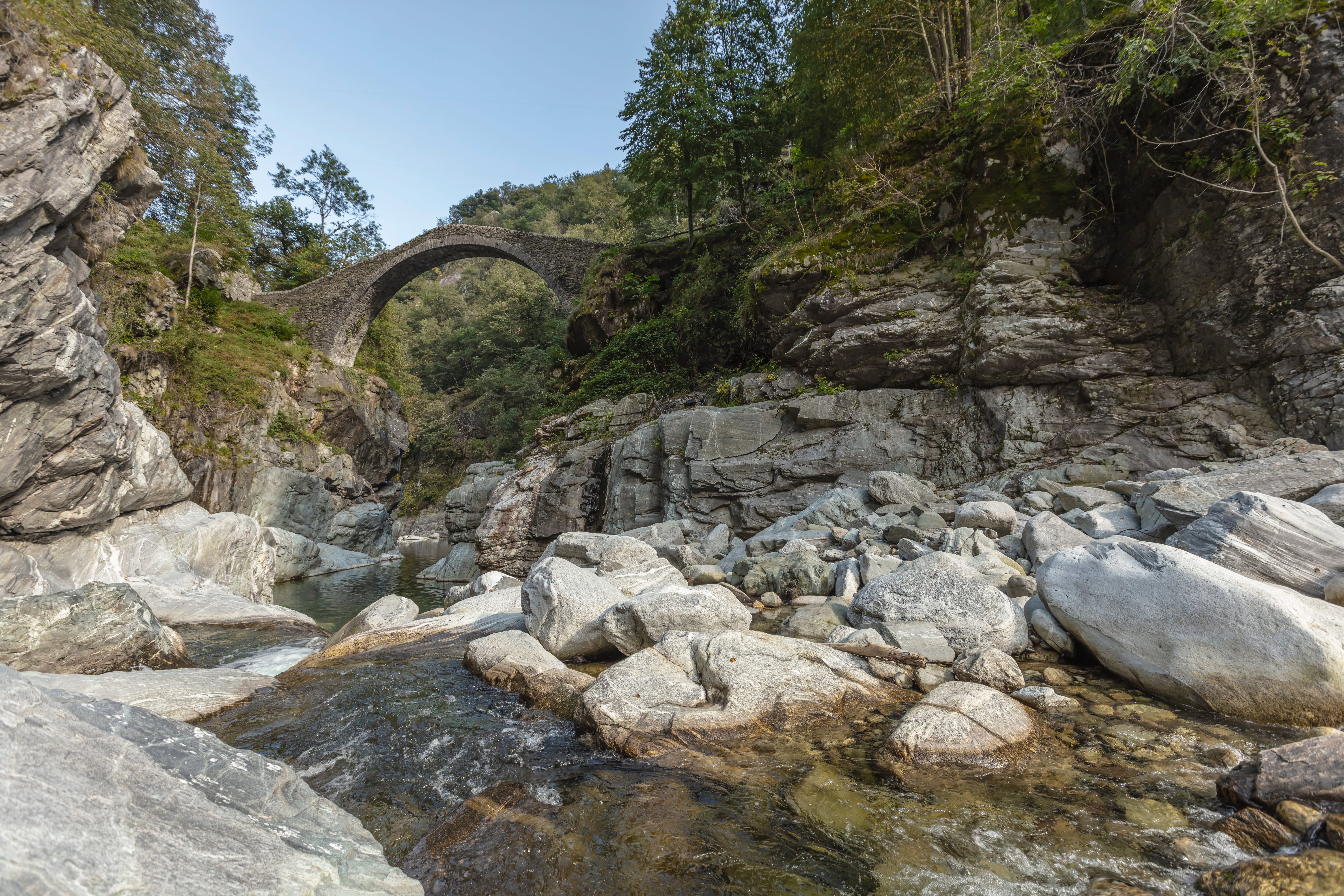 Ancient stone arch bridge spanning across a rocky gorge with large boulders and shallow stream below, surrounded by green trees.