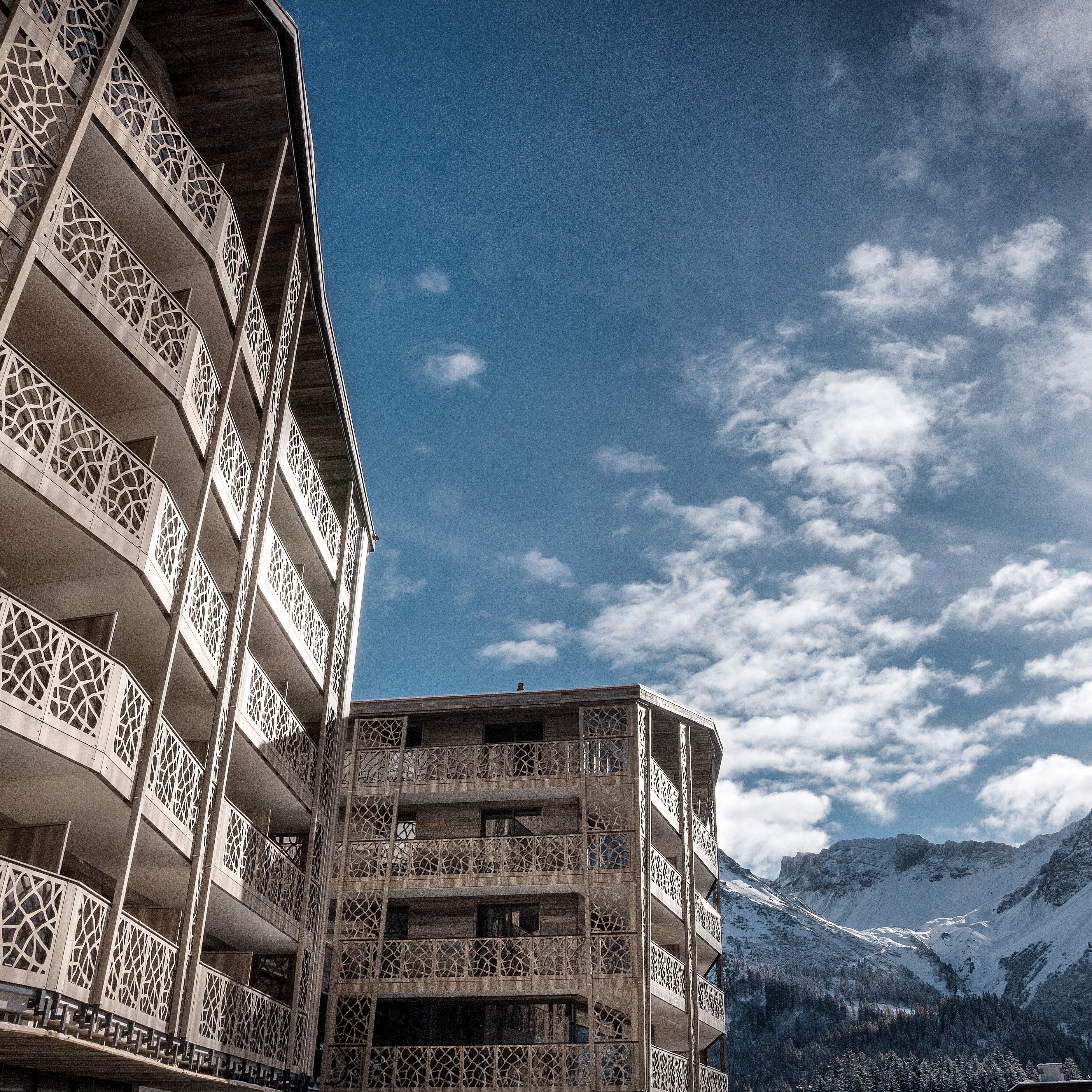 Moderne Gebäude mit dekorativen Balkonen vor schneebedeckten Bergen und blauem Himmel mit weissen Wolken.