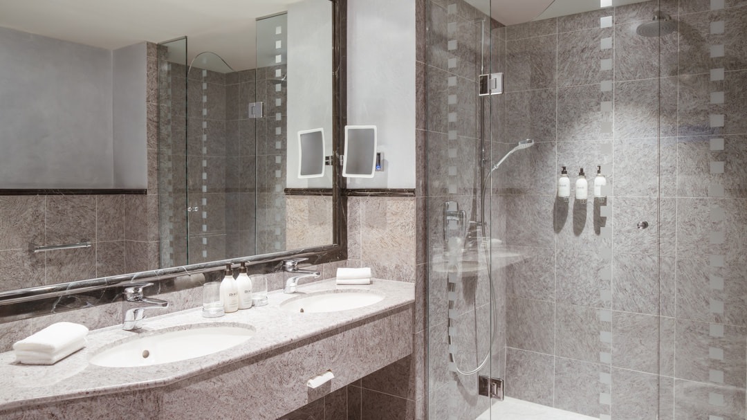 Elegantes Badezimmer mit großem Spiegel, zwei Waschbecken aus Stein, Glasdusche mit Regenduschkopf und hochwertigen Pflegeprodukten auf Natursteinfliesen.