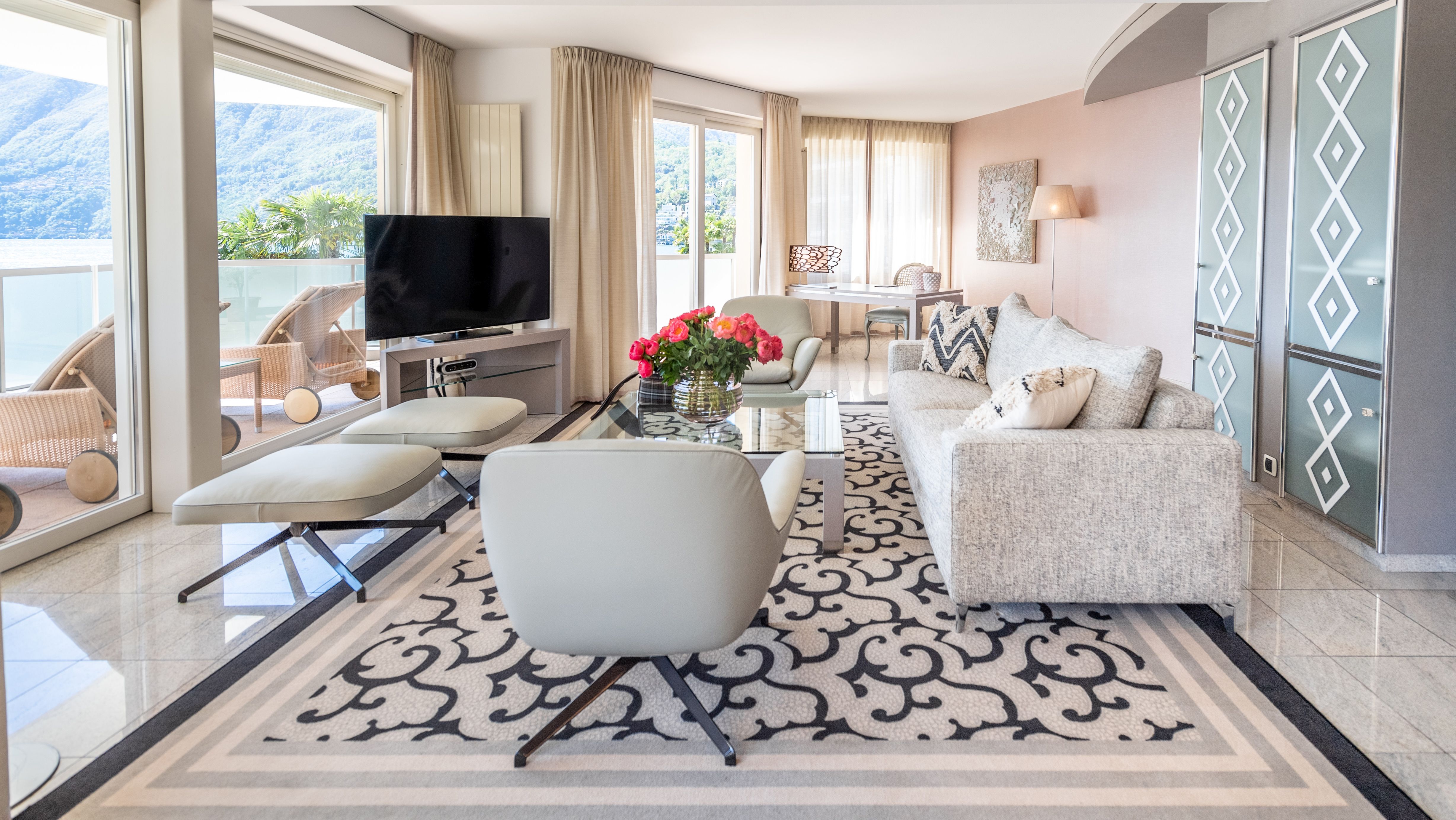 Modernes Wohnzimmer mit hellen Möbeln, gemustertem Teppich, Seeblick durch grosse Fenster und rosa Akzentwand.