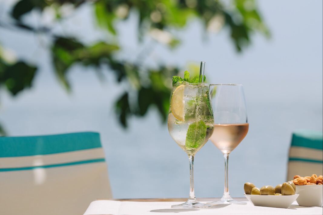 Zwei Weingläser auf einem Tisch, eines mit Cocktail garniert mit Minze und Limette, eines mit Roséwein, Snacks daneben.
