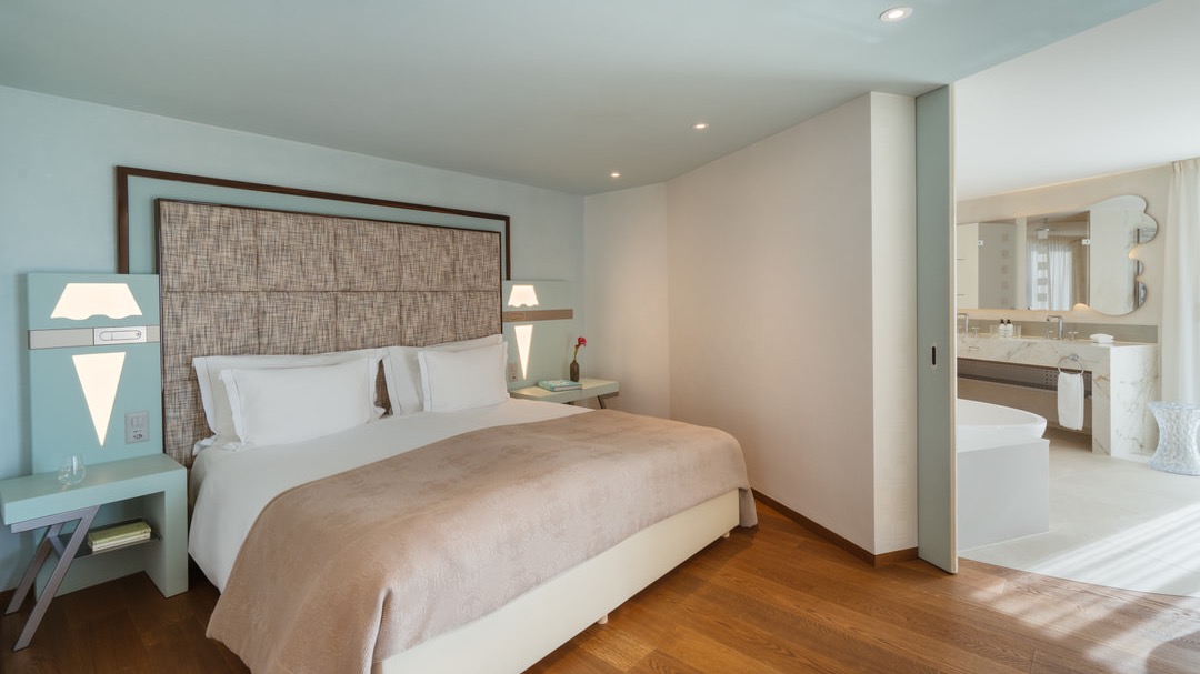 Stilvolles Hotelzimmer mit modernem Doppelbett, hellblauem Interieur, weichem Holzboden und offenem Übergang zum luxuriösen Badezimmer mit freistehender Badewanne und Doppelwaschtisch.