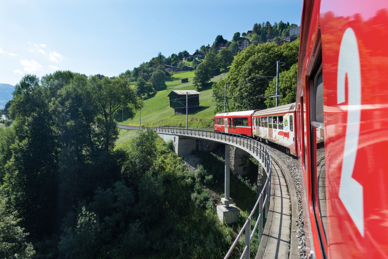 Red and white train crossing a curved bridge through green hillside with trees, buildings, and Swiss flag visible.