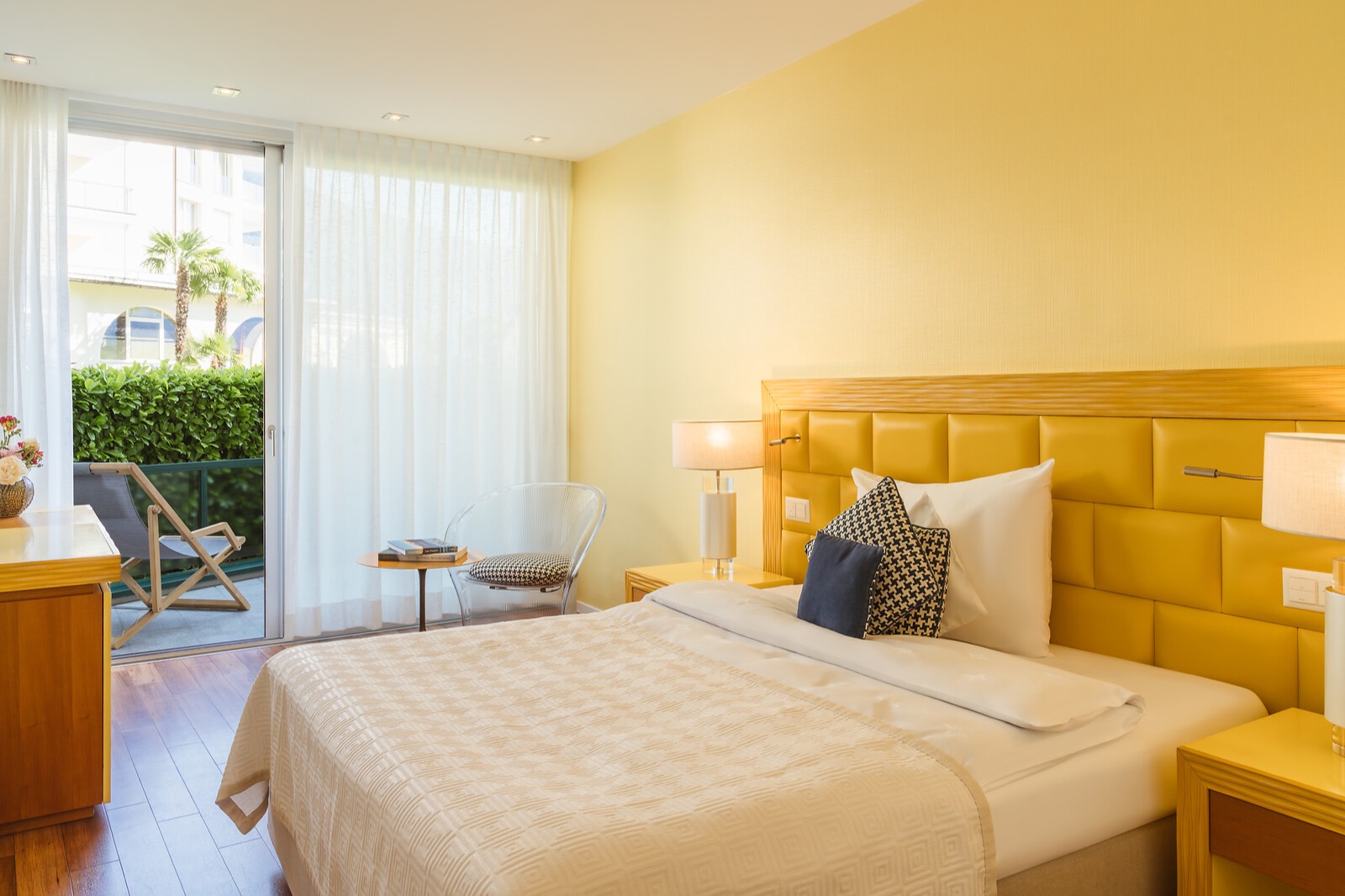 Modern hotel bedroom with yellow accent wall, upholstered headboard, white bedding, and balcony access with sheer curtains.