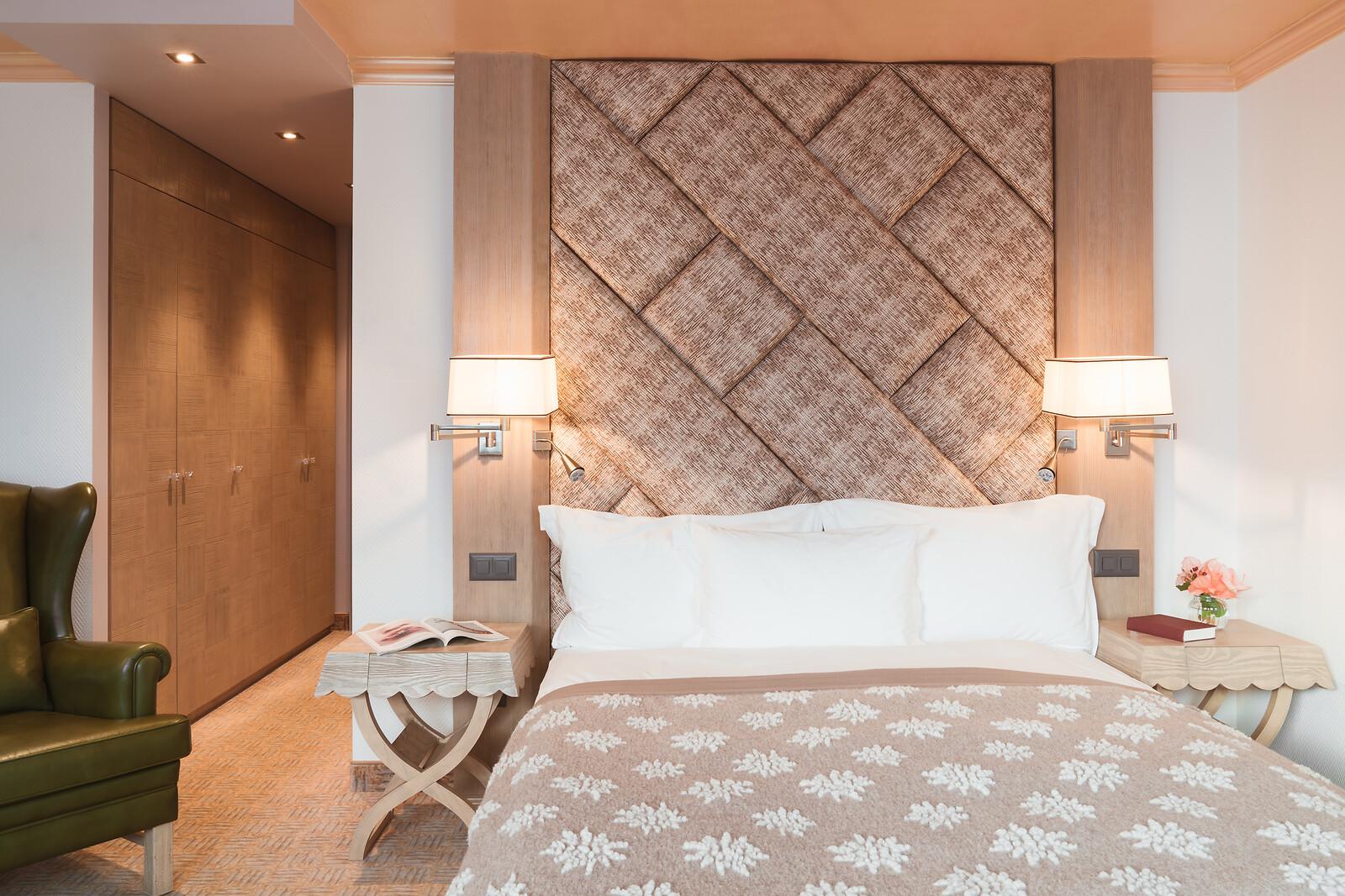 Elegantes Hotelzimmer mit grossem Doppelbett, geflochtenem Kopfteil als Blickfang, weissen Kissen und beiger Tagesdecke mit Blumenmotiv, ergänzt durch Holznachttische, warme Wandleuchten, Einbauschränke und einen grünen Ledersessel.