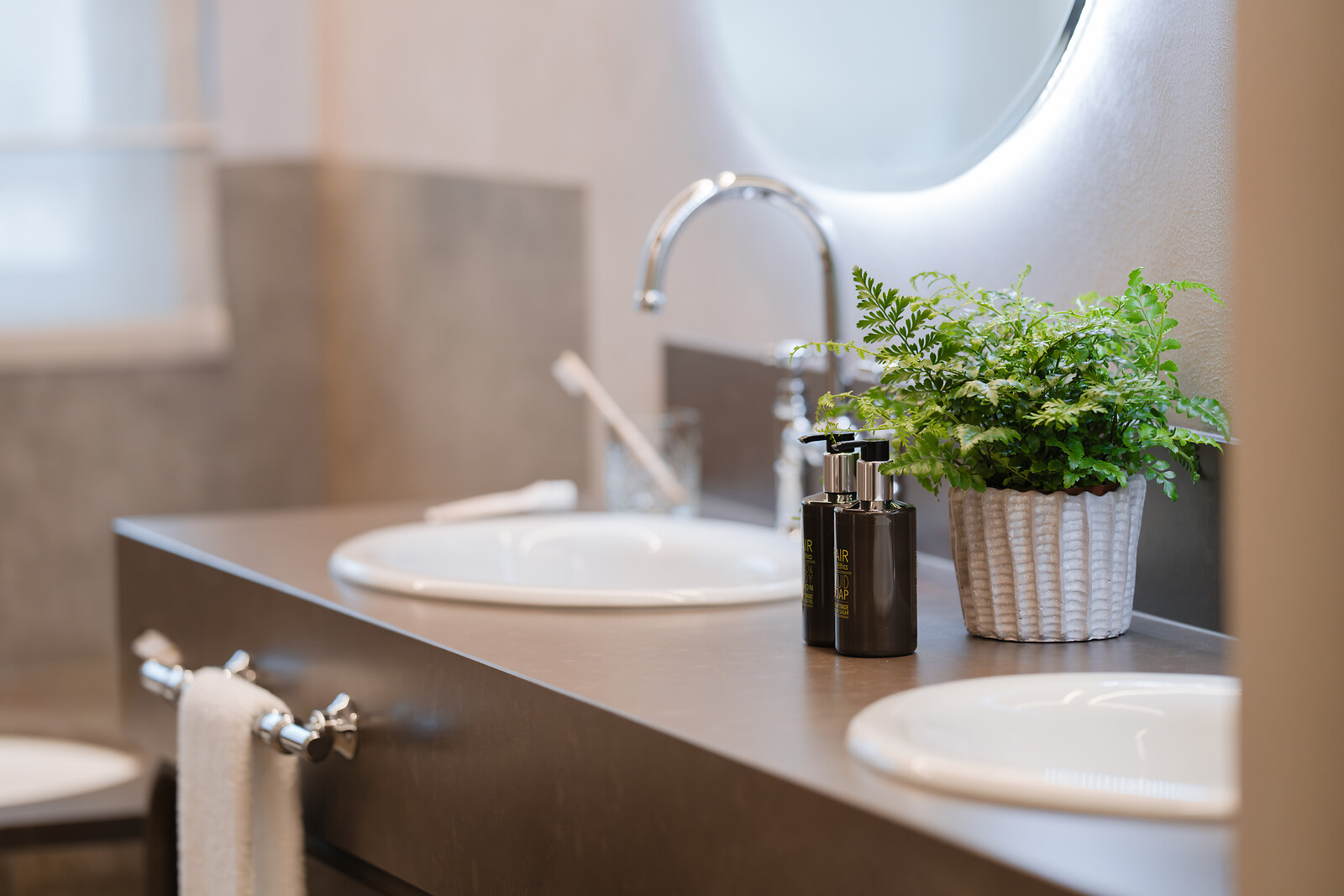 Modernes Badezimmer mit Doppelwaschbecken, eleganten Armaturen, Pflegeprodukten und dekorativer Pflanze auf der Ablage.