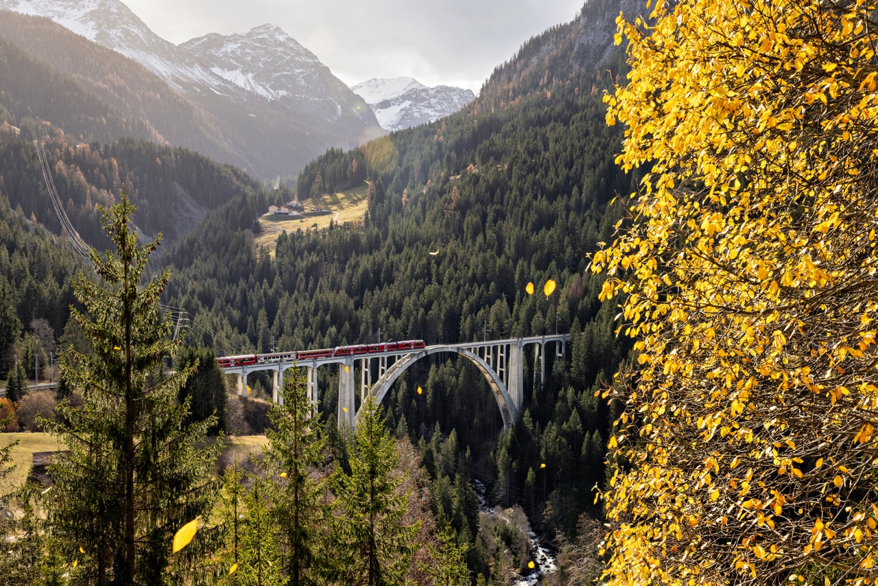 Red train crossing a curved stone viaduct through an alpine valley with autumn foliage and snow-capped mountains.