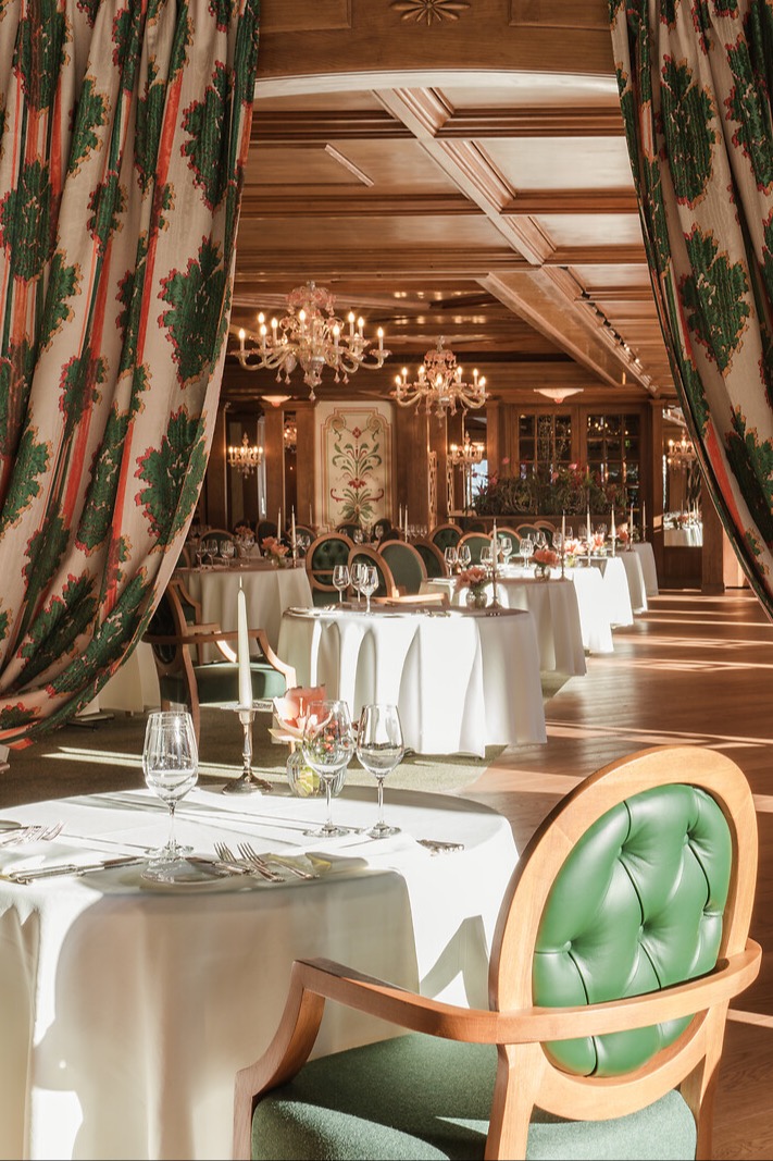 Elegant restaurant dining room with white-clothed tables, green upholstered chairs, crystal chandeliers, and decorative curtains.