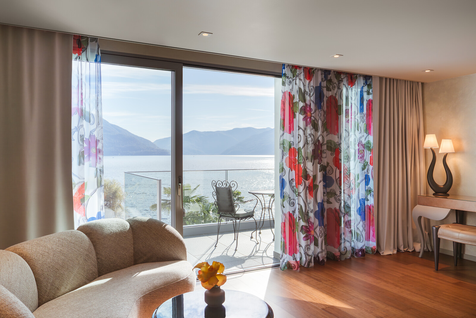 Helles Wohnzimmer mit Panoramafenster, floral gemusterten Vorhängen, beiger Designercouch, Glastisch mit gelber Blume und Balkon mit Blick auf See und Berge.