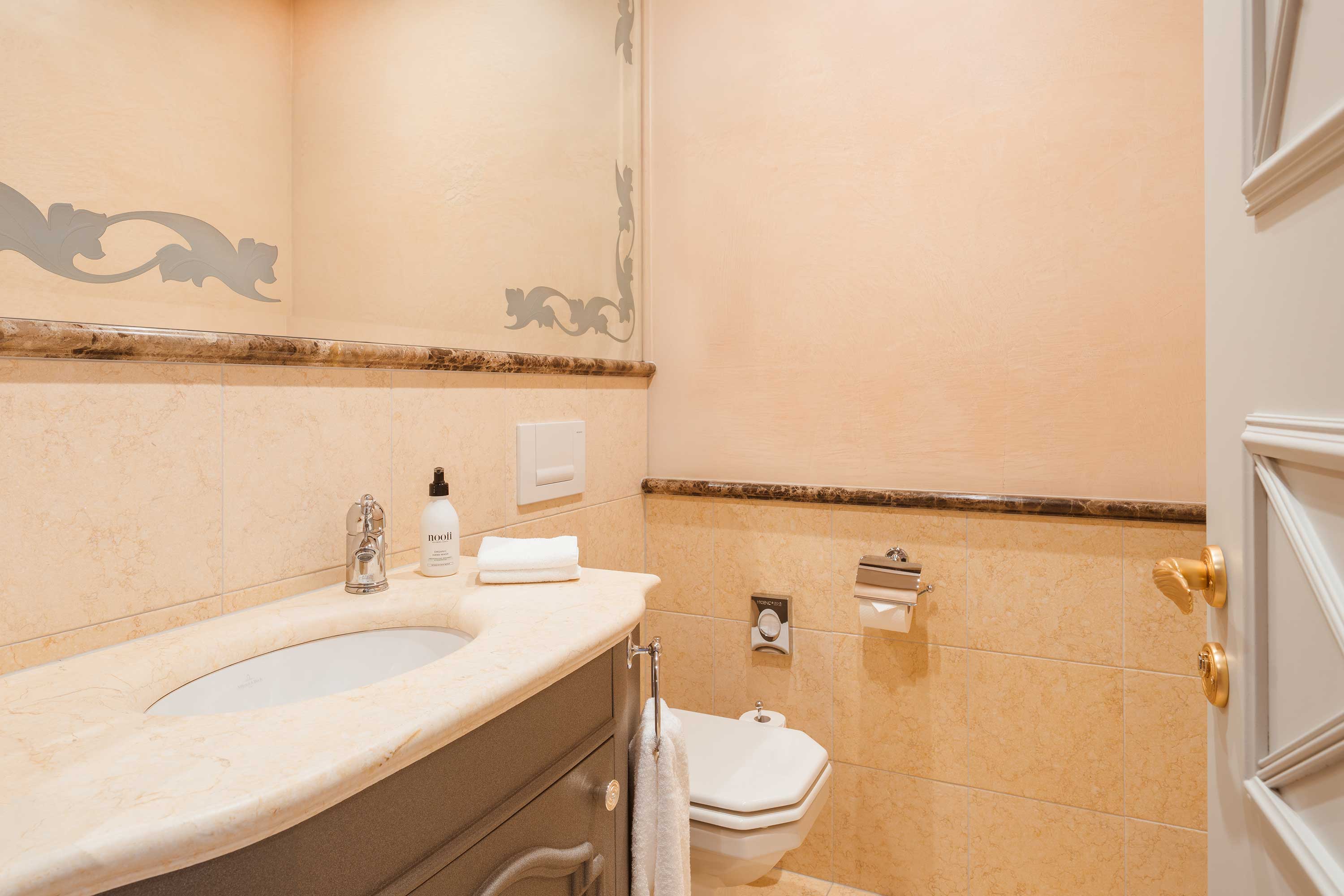 Elegant bathroom with peach walls, beige marble countertop, white sink, toilet, decorative gray stencil border, and gold fixtures.