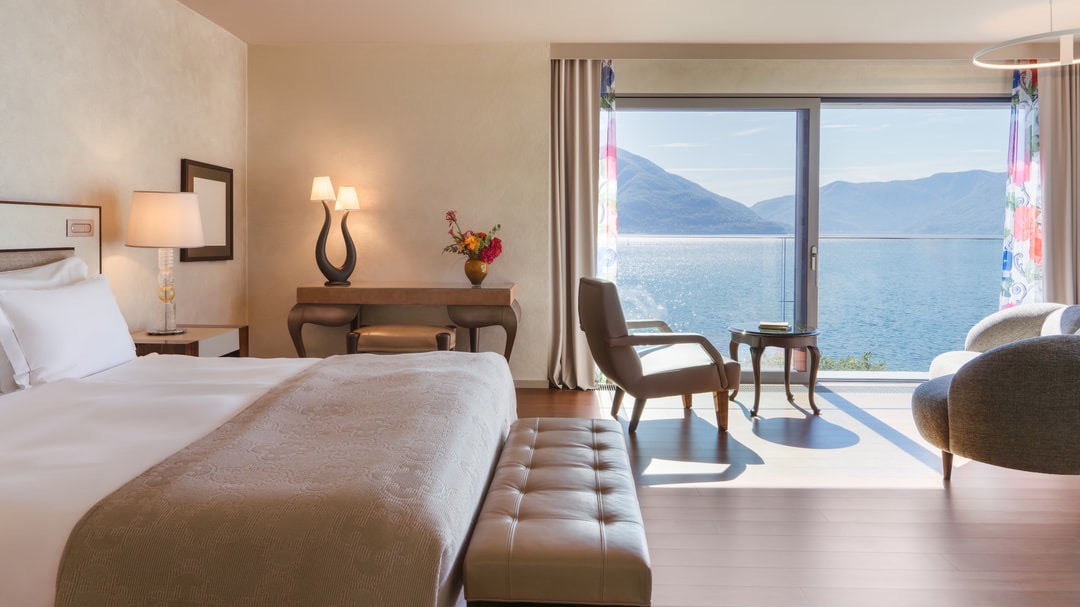 Helles Schlafzimmer mit Kingsize-Bett, eleganter Sitzgruppe, großem Fenster mit Seeblick, floralen Vorhängen und stilvollen Möbeln auf hellem Holzboden.