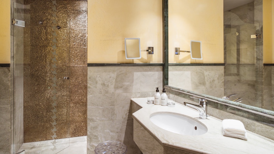 Modernes Badezimmer mit goldener Mosaikdusche, großem Spiegel, Waschbecken mit Pflegeprodukten und heller Wandgestaltung.