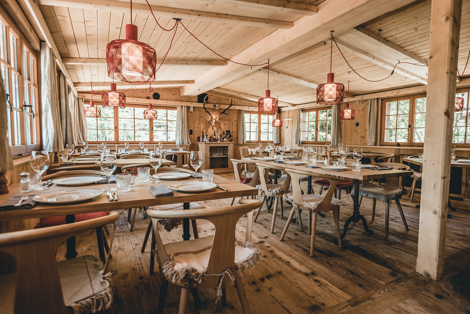 Rustic Alpine restaurant interior with wooden tables and chairs, exposed beams, large windows and red pendant lights, creating a warm and welcoming atmosphere.