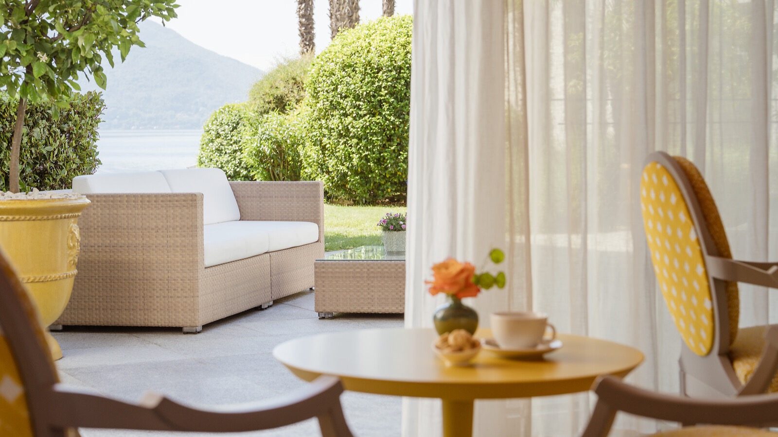 Elegante Terrasse mit beigefarbenen Rattanmöbeln, weissen Vorhängen und Blick auf grünen Garten und See