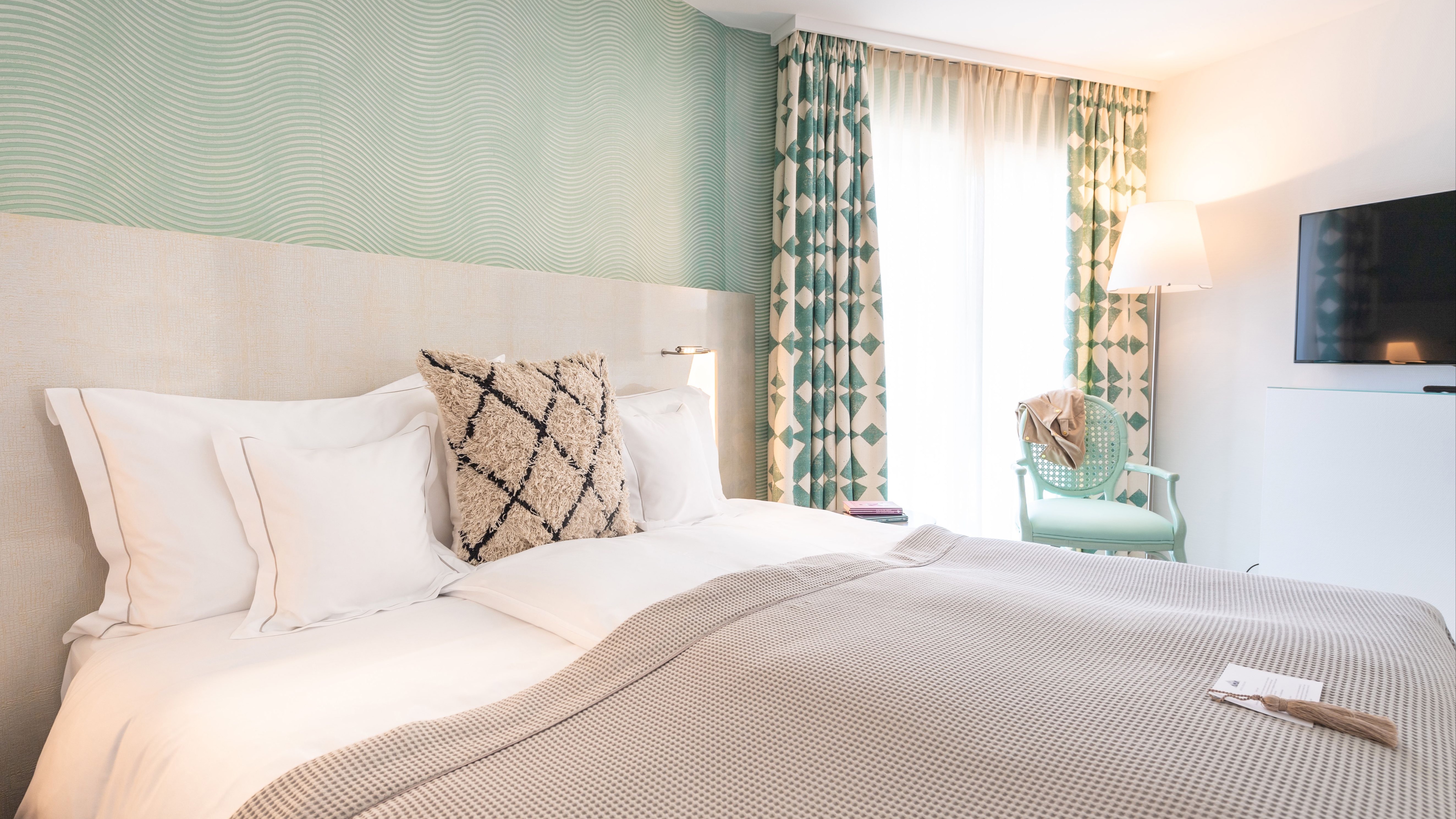 Modern bedroom with white bedding, green wavy textured accent wall, geometric patterned curtains, and mint green chair.