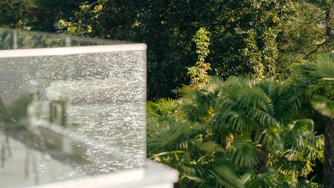 Close-up of a glass balcony with sunlight reflections, palm trees in the foreground, and dense forest backdrop – nature meets luxury.