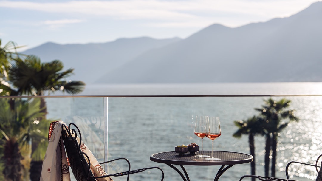 Romantic balcony with lake view, palm trees and mountains, round metal table set with two glasses of rosé wine, small snacks, and stylish garden chairs.