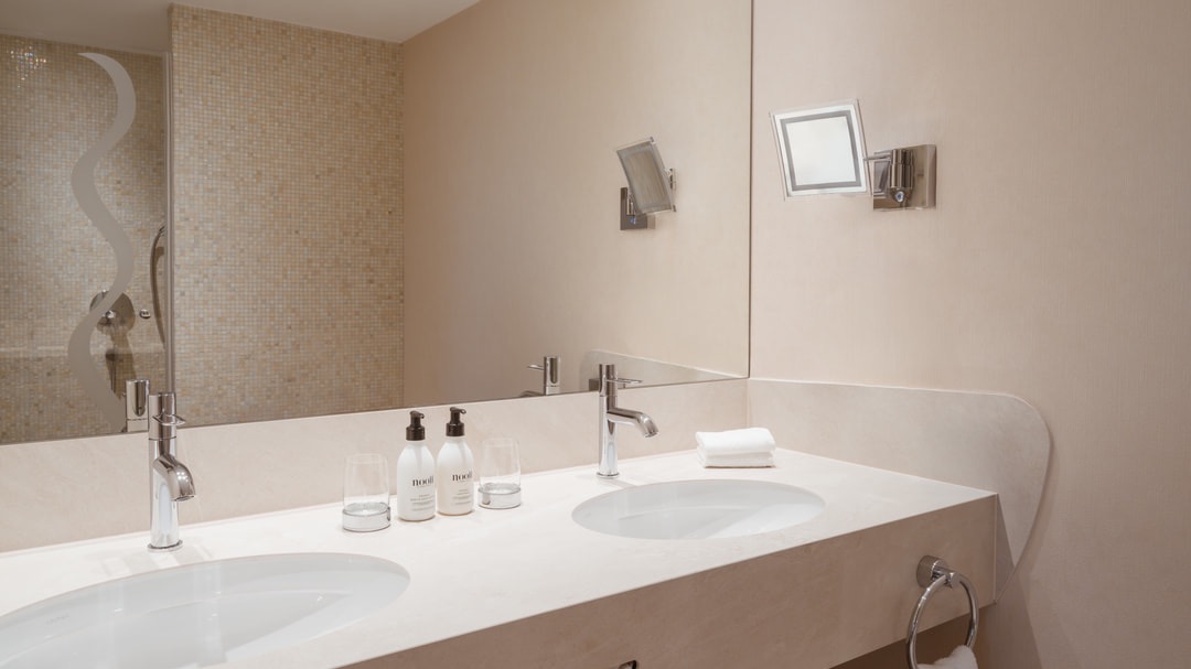 Modernes Badezimmer mit Doppelwaschtisch, großen Wandspiegeln, Pflegeprodukten, Mosaikdusche im Hintergrund und minimalistischer Beleuchtung.