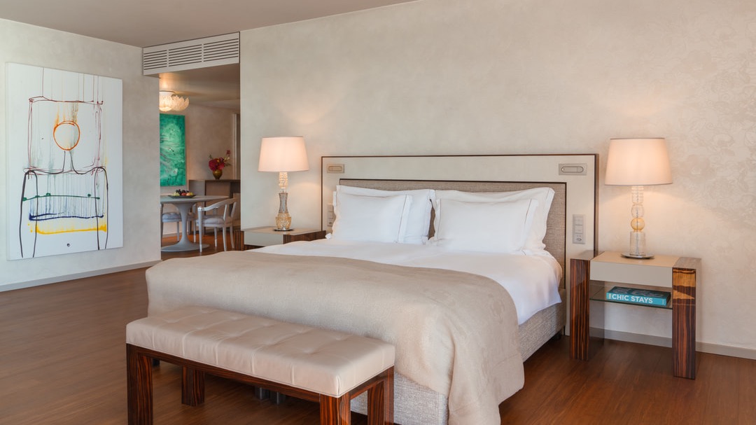 Modernes Schlafzimmer mit Doppelbett, beigem Bettüberwurf, Nachttischen mit Designerlampen, Wandkunst und Blick in den offenen Essbereich.