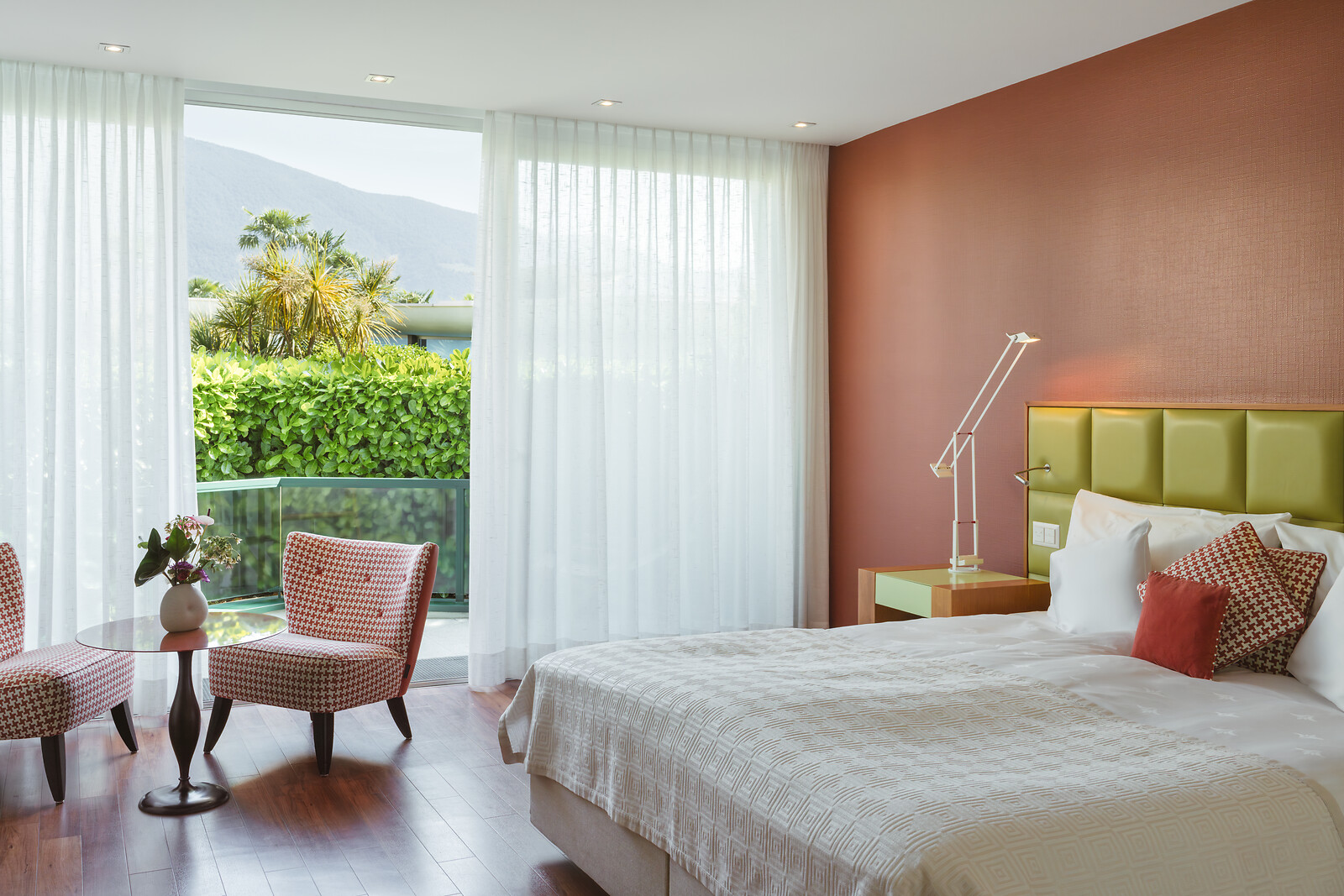 Modern bedroom with terracotta accent wall, yellow-green headboard, white curtains, patterned chairs, and balcony view.