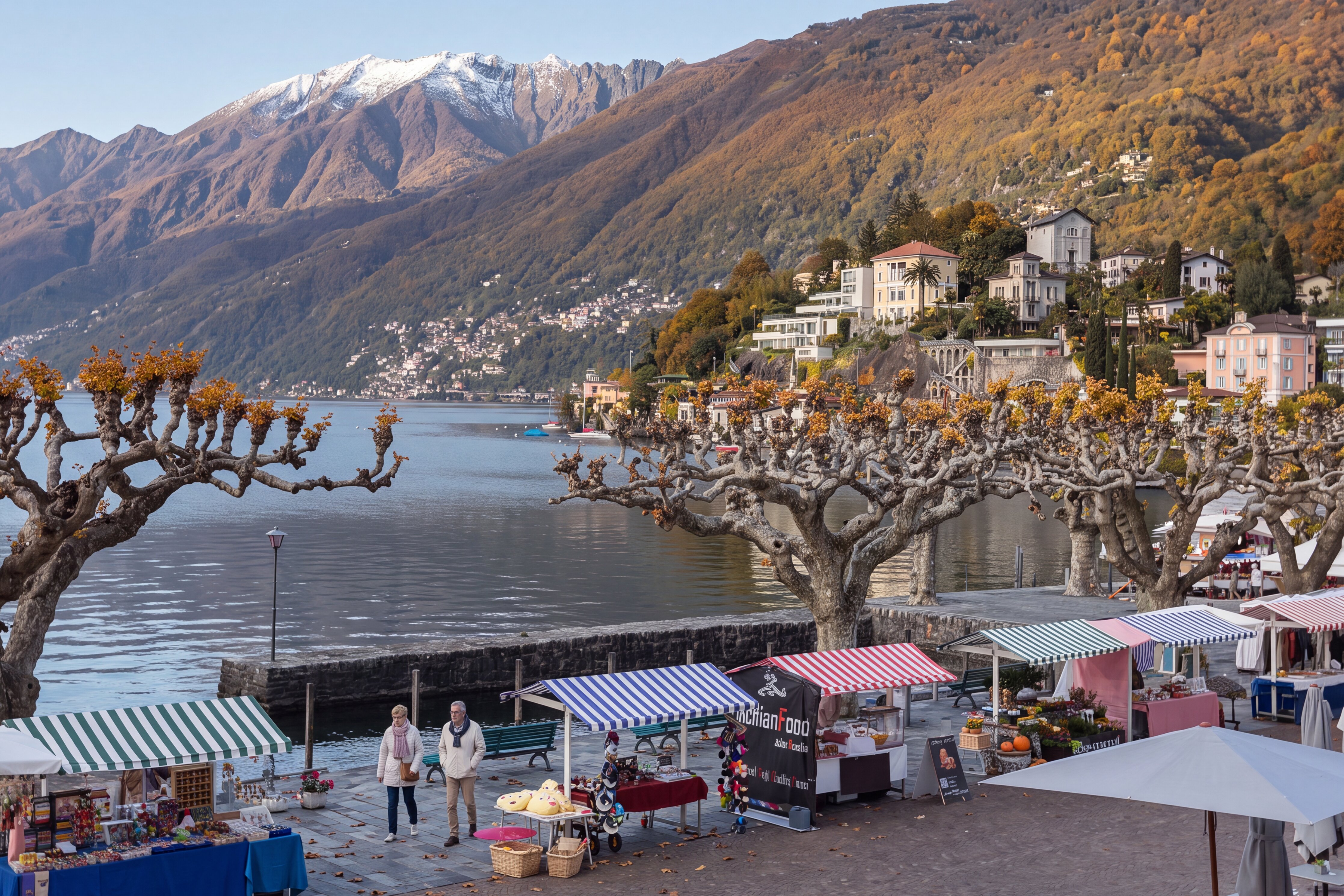 Herbstlicher Markt am Seeufer von Ascona mit bunt gestreiften Marktständen, kahlen Platanen und einem spazierenden Paar. Im Hintergrund liegen der Lago Maggiore, historische Villen und bewaldete Berge mit schneebedeckten Gipfeln.